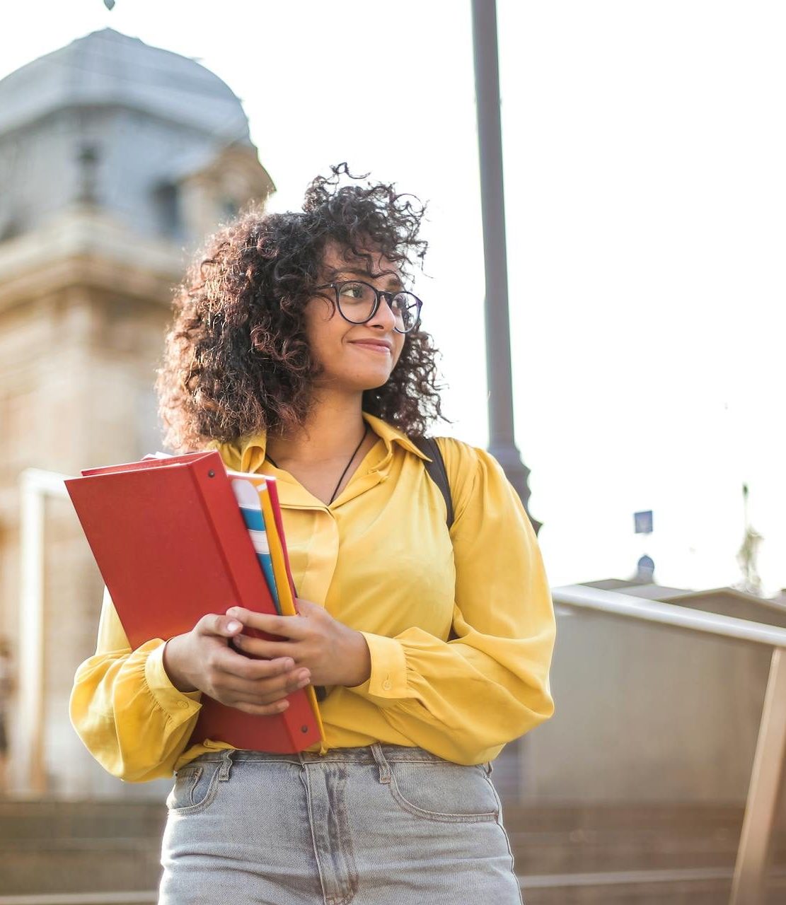 Lifestyle Foto Mädchen auf Campus Mädchen mit braunen Locken, Brille und gelber Bluse. Im Hintergrund sieht man den Campus einer Uni.