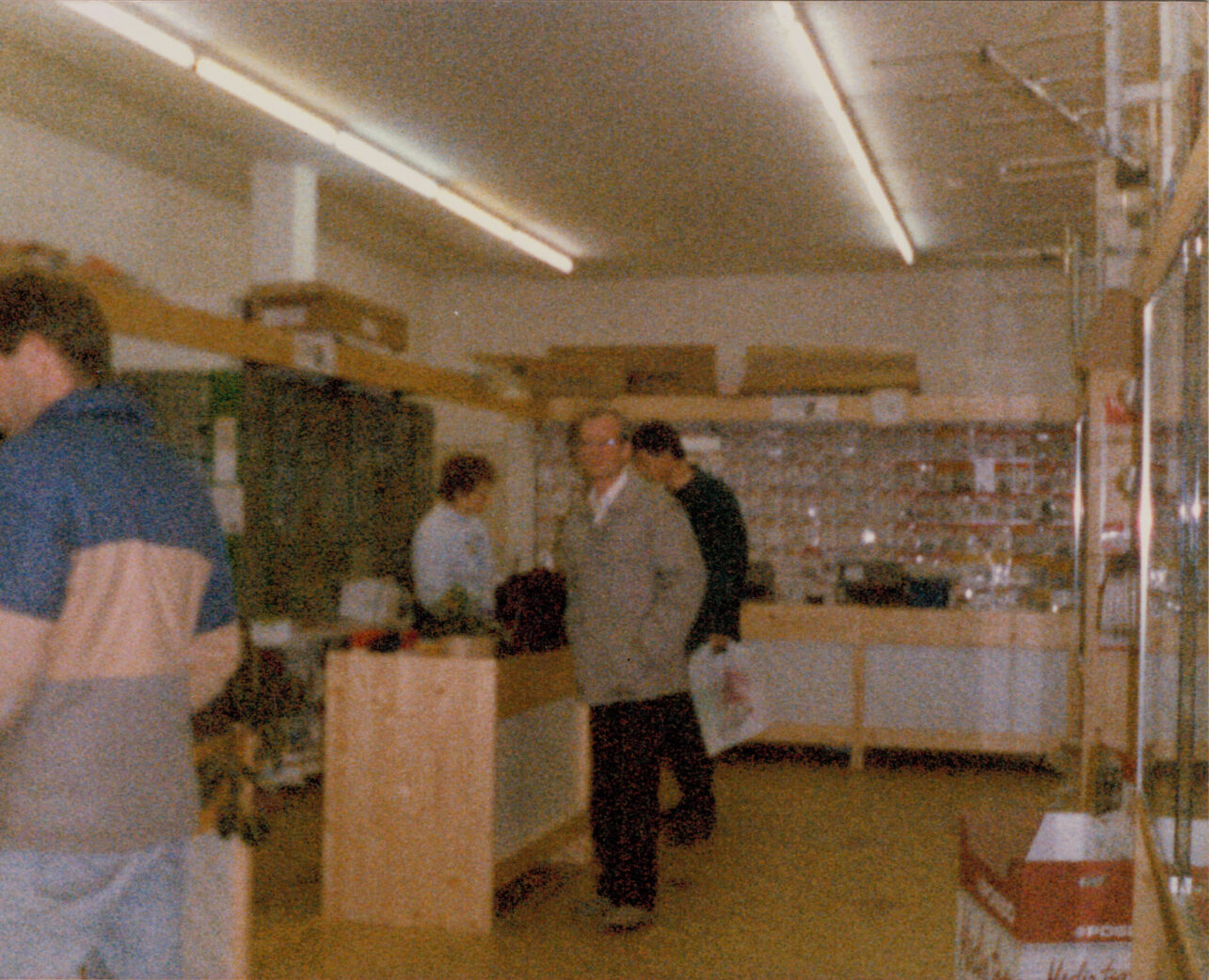 Das Bild zeigt die Eröffnung des Ladens in der Kasinostraße im Jahr 1987. Im Vordergrund ist eine hölzerne Ladentheke zu sehen, während sich im Hintergrund mehrere Besucher versammelt haben. Die Aufnahme fängt die besondere Atmosphäre dieses historischen Moments ein.