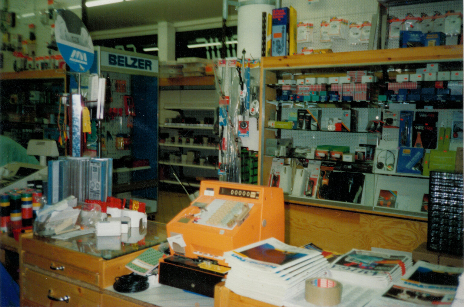 Das Bild zeigt ein Ladengeschäft der 1990er Jahre mit einer alten Kasse auf der Ladentheke. Daneben liegen Lamiga-Zeitschriften aus, die einen nostalgischen Einblick in das damalige Sortiment und die Technik dieser Zeit bieten.