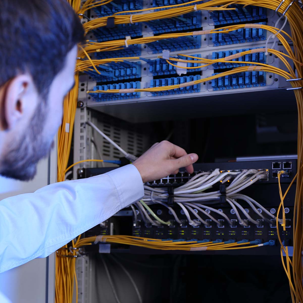 Lifestyle Foto IT-Ingenieur in Serverraum_2 Das Bild zeigt einen männlichen IT-Ingenieur, der in einem Serverraum arbeitet und für die Netzwerksicherheit sorgt. Er überprüft sorgfältig die angeschlossenen Kabel an einem Server, was auf eine präzise und verantwortungsvolle Arbeitsweise in der IT-Infrastruktur hinweist. Der Raum ist mit zahlreichen Servern und technischen Geräten ausgestattet, die eine professionelle Arbeitsatmosphäre vermitteln.