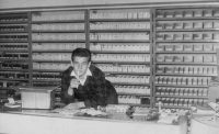 Lutz Zimmermann an Ladentheke 1962. Im Hintergrund sieht man Kistchen mit Ersatzteilen in schwarz und weiß.