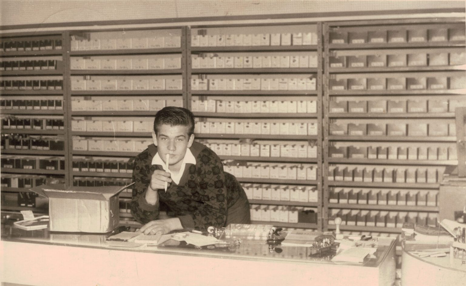 Das Bild zeigt Lutz Zimmermann im Jahr 1962 an der Ladentheke. Im Hintergrund sind kleine Kisten mit Ersatzteilen zu sehen. Die Aufnahme vermittelt einen nostalgischen Einblick in das Geschäftsumfeld der damaligen Zeit.