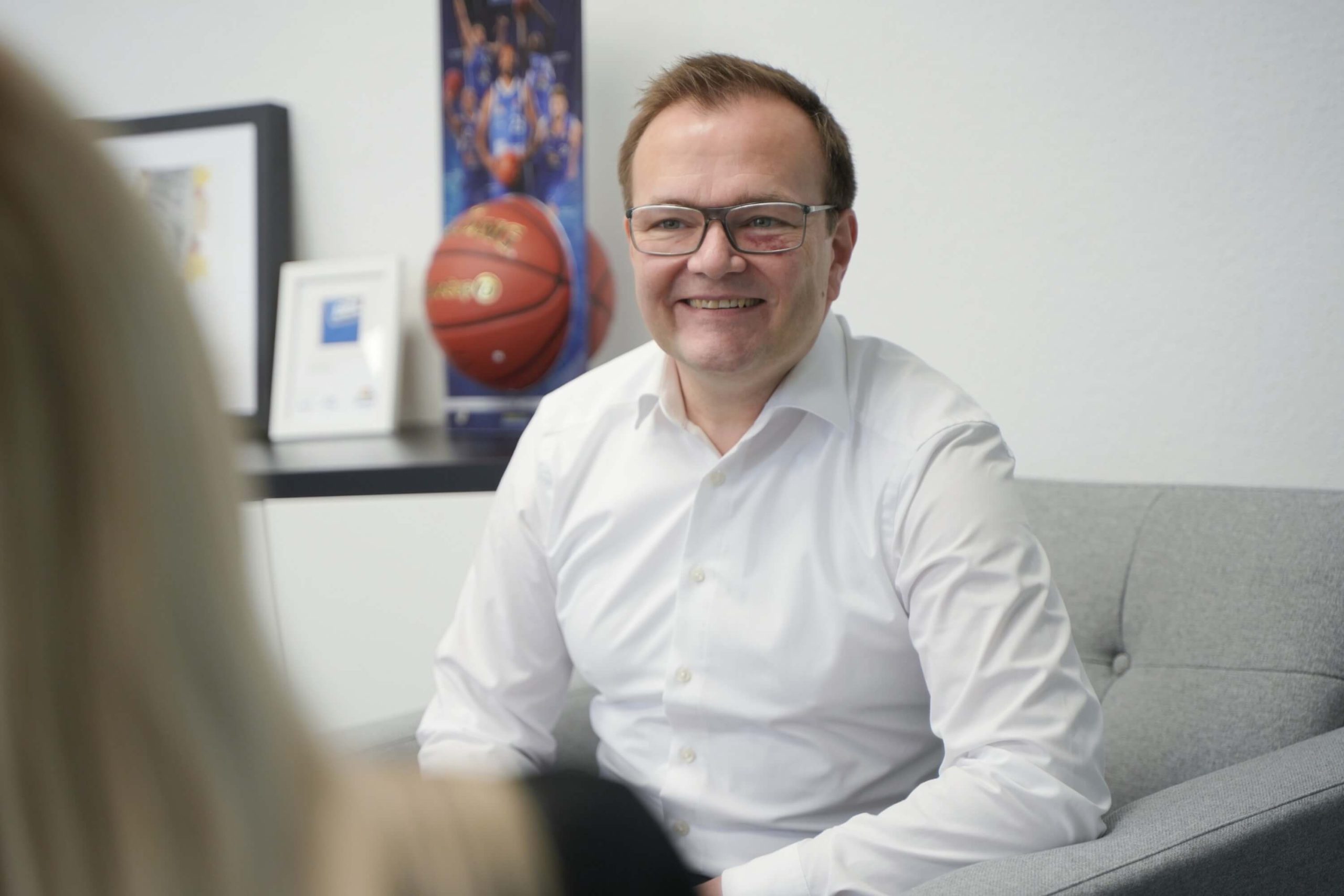 Vorstellungsgespräch Situation MZ CEO Michael Zimmermann von Rensch IT-Consult, ein lächelnder Mann mit Brille in einem weißen Hemd, sitzt auf einem grauen Sofa während eines Vorstellungsgesprächs mit einer Bewerberin im schwarzen Blazer. Im Hintergrund sind ein Basketball und eine Urkunde sichtbar.