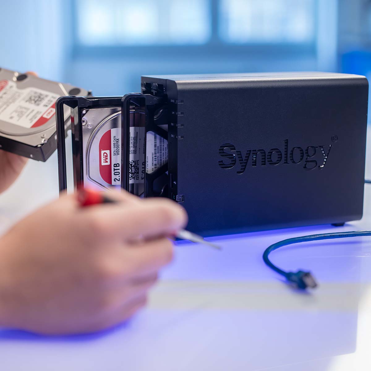 Situationsbild NAS HDDs Tausch Das Bild zeigt einen Mitarbeiter, der eine NAS-HDD (Network-Attached Storage Hard Drive) austauscht. Der Mitarbeiter arbeitet sorgfältig und präzise, um die Festplatte zu entfernen und durch eine neue zu ersetzen. Die Szene vermittelt einen technischen Arbeitsprozess in einer professionellen IT-Umgebung, bei dem auf Datenintegrität und Netzwerkoptimierung geachtet wird.