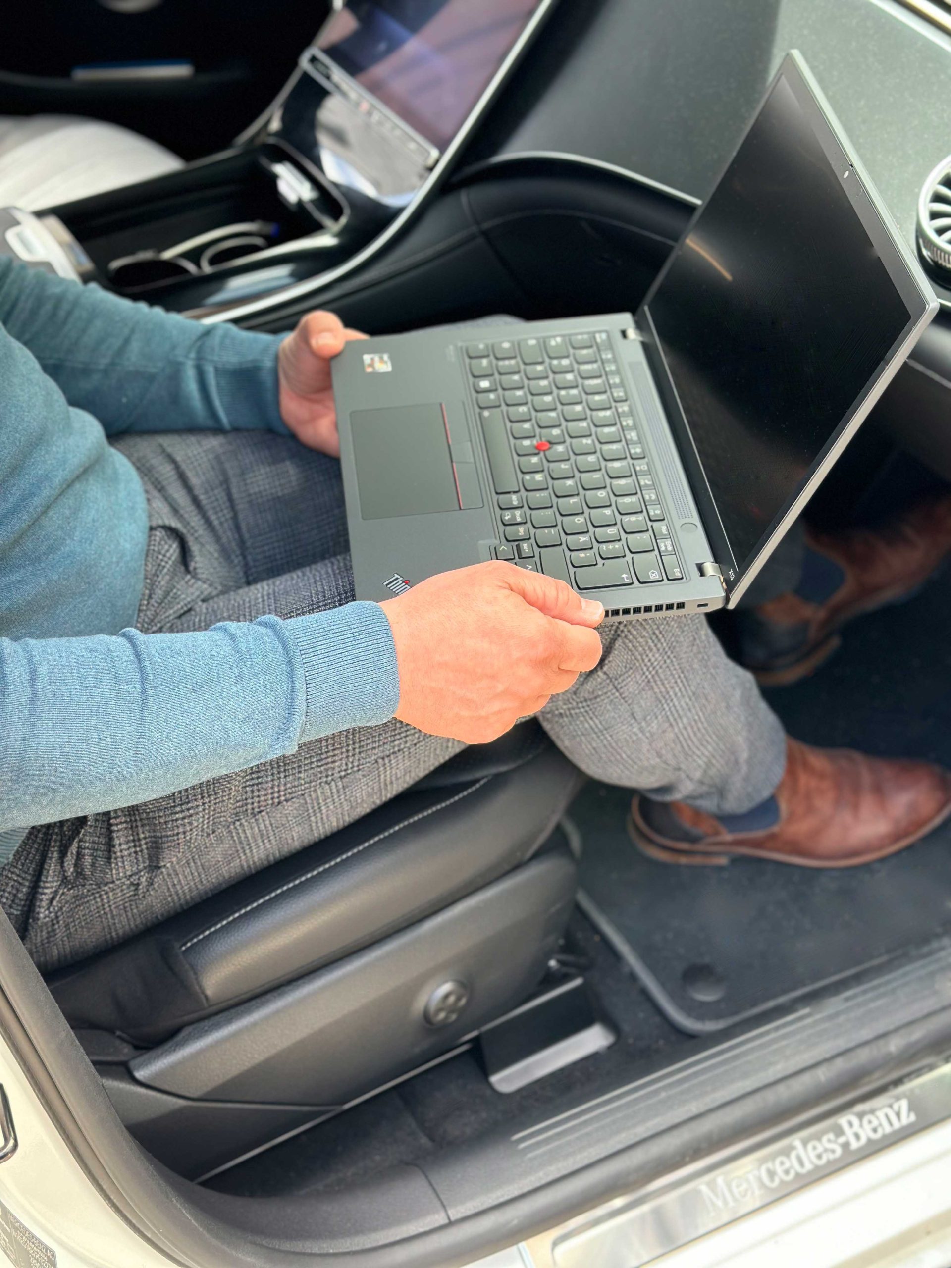 Ein Mann sitzt im Auto mit einem Laptop auf dem Schoß, während er konzentriert darauf arbeitet. Die Szene vermittelt Flexibilität und den Einsatz moderner Technologie, auch unterwegs.