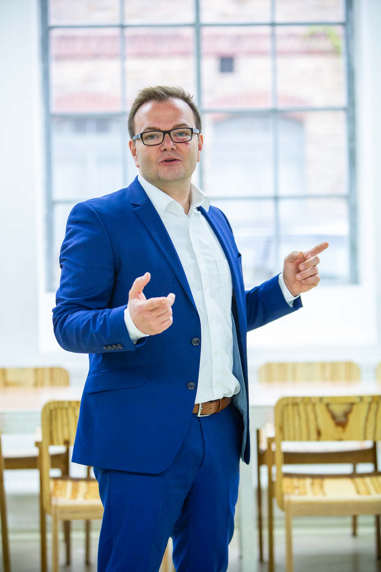 M. Zimmermann steht mit einer freundlichen Handbewegung, die eine einladende Geste ausdrückt. Er trägt einen blauen Anzug und ein weißes Hemd, was ihm ein professionelles und zugleich sympathisches Auftreten verleiht.