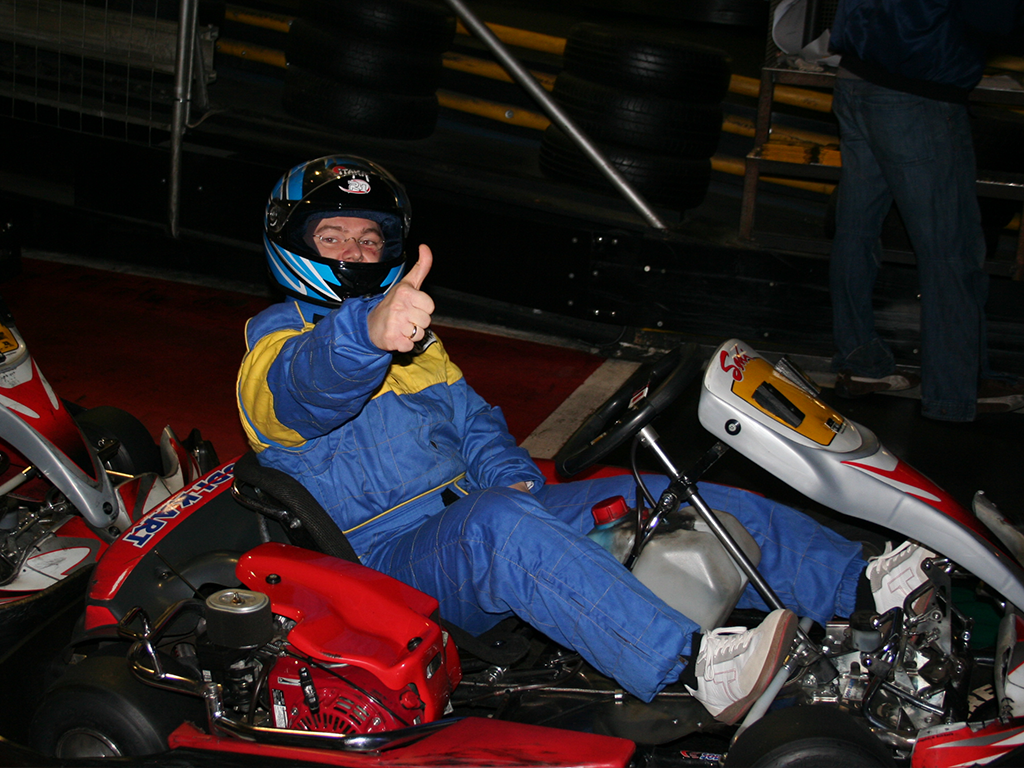 Der CEO des Unternehmens fährt beim Kartfahren während der Firmenweihnachtsfeier. Er trägt einen blau-gelben Kartanzug und einen Helm, während er mit dem Daumen nach oben auf die Kamera zeigt. Die Szene vermittelt eine entspannte, fröhliche Atmosphäre und zeigt den CEO in einer lockeren, teamorientierten Situation.
