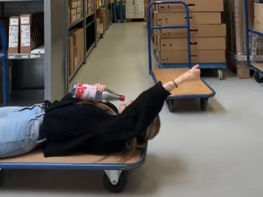 ine Mitarbeiterin liegt singend auf dem Rücken auf einem Rollbrett im Lager und streckt ihren linken Arm nach hinten, als wäre sie ein Superheld. In ihrer rechten Hand hält sie eine Wasserflasche wie ein Mikrofon und bringt ihre Leidenschaft und Energie zum Ausdruck, während sie mit einer fröhlichen und humorvollen Haltung das Lager in eine kleine Bühne verwandelt.