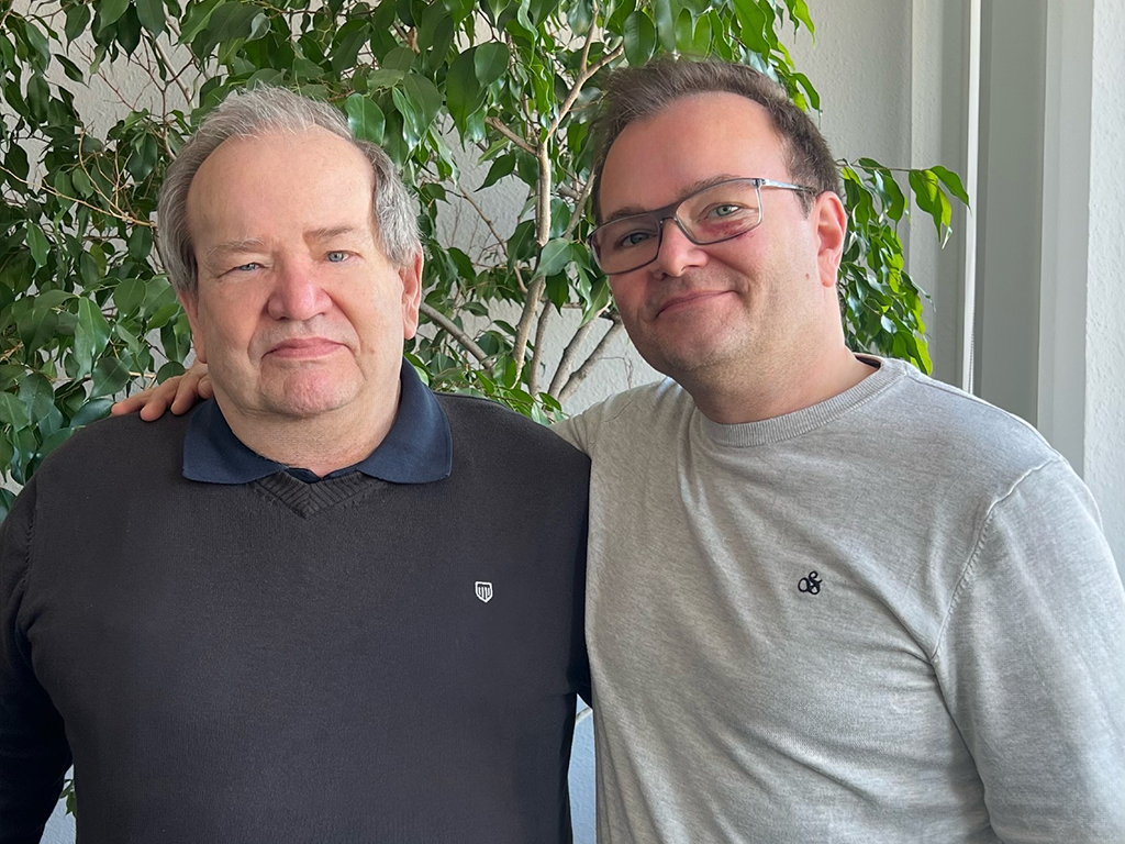 Michael und Lutz Zimmermann stehen vor einer Pflanze. Michael hat seinen rechten Arm freundschaftlich um Lutz gelegt, was eine enge und vertraute Beziehung zwischen den beiden zeigt. Die Szene strahlt eine warme und kollegiale Atmosphäre aus.