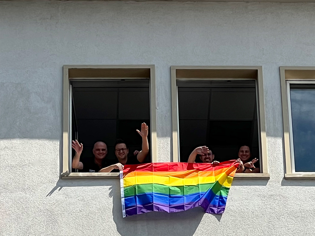 Vier Mitarbeiter halten eine farbenfrohe Pride-Flagge aus dem Fenster und genießen einen fröhlichen Moment zusammen. Lächelnd und voller Energie zeigen sie ihre Unterstützung für die LGBTQ+-Community und feiern Vielfalt und Zusammenhalt in einer offenen, positiven Atmosphäre.
