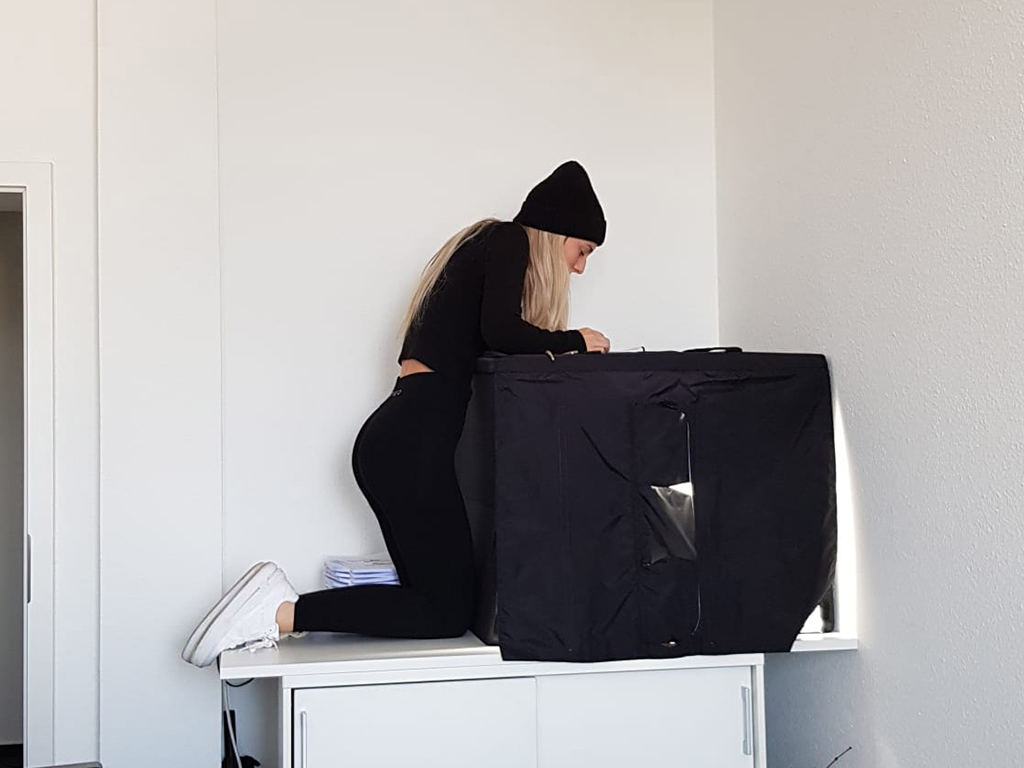 Vanessa macht Produktfotos an einer schwarzen Fotobox. Sie trägt eine schwarze Mütze und weiße Schuhe, während sie auf einem Tisch kniet, um von oben in die Box zu schauen. Ihre konzentrierte Haltung und die präzise Arbeit vermitteln eine professionelle Atmosphäre für das Fotografie-Shooting.